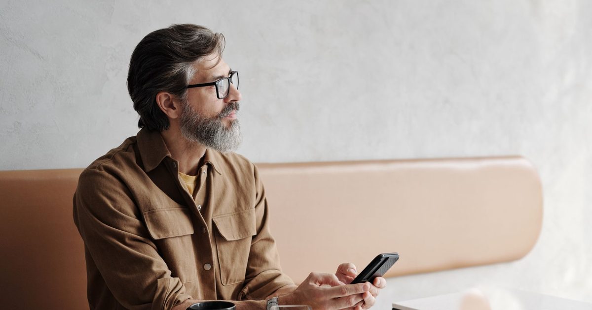 Uomo con la barba in un bar con il telefono in mano, lo sguardo pensieroso