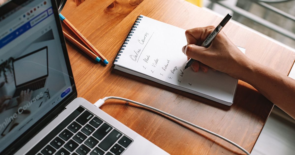 Mano che scrive appunti su un quaderno accanto a un portatile aperto